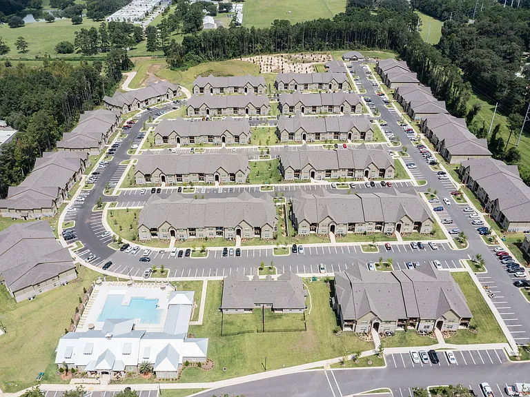 Scenic aerial view of a sprawling apartment community with neatly arranged buildings, ample parking, and lush green surroundings, featuring a luxurious pool area for residents.