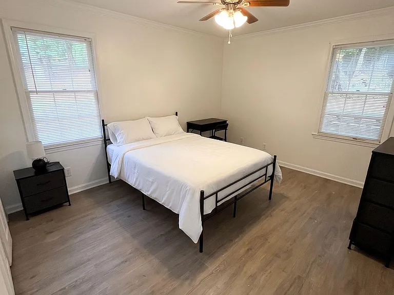 Furnished bedroom with a full-size bed, desk area, and two windows for natural light plus a ceiling fan overhead.