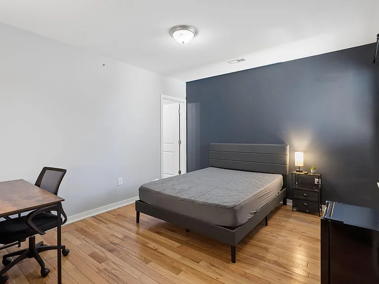 Furnished bedroom with a comfortable bed, nightstand lighting, hardwood floors, and a dedicated desk/work area.