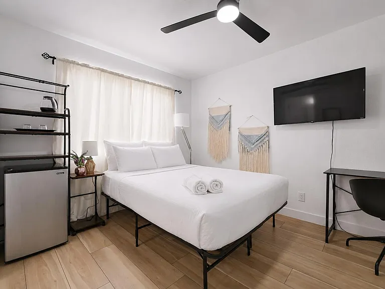 Well-appointed bedroom with a ceiling fan, wall-mounted TV, work desk, and an in-room mini fridge plus open shelving.
