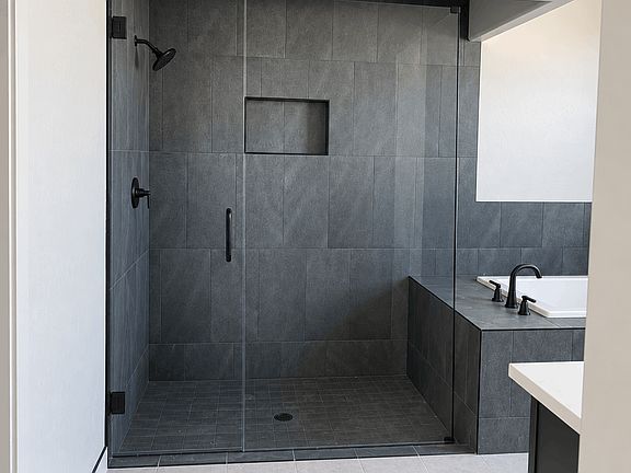A walk-in shower with dark gray tile walls and a built-in recessed shelf, featuring a glass door and