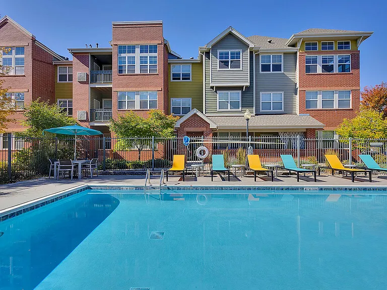 Charming apartment building with a vibrant outdoor pool area, colorful lounge chairs, and lush green landscaping perfect for relaxation and social gatherings.