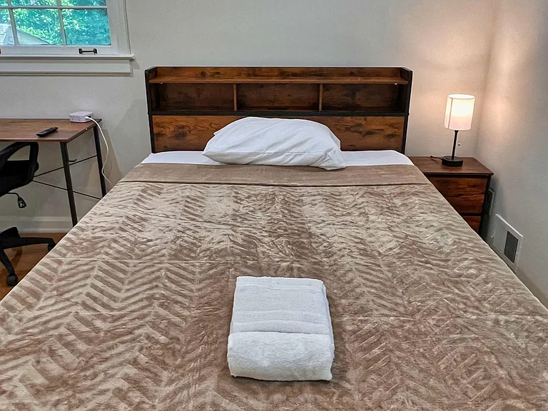 Furnished bedroom with a full bed, nightstand lighting, and a dedicated desk workspace plus a window for natural light.