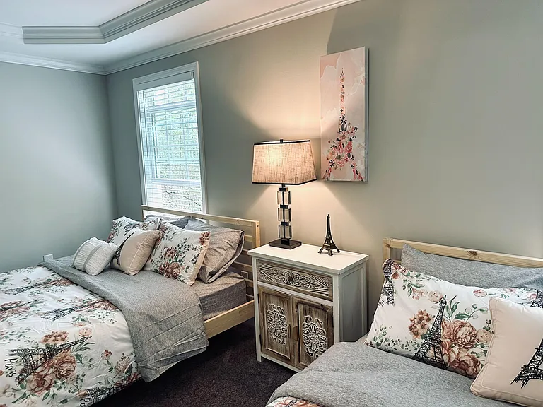 Furnished bedroom with two twin beds, a bedside cabinet and lamp, and a window with blinds for natural light.