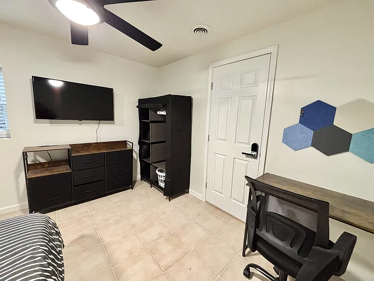 Spacious bedroom with full sized bed, mounted smart TV, desk, bulletin board and standing armoire.