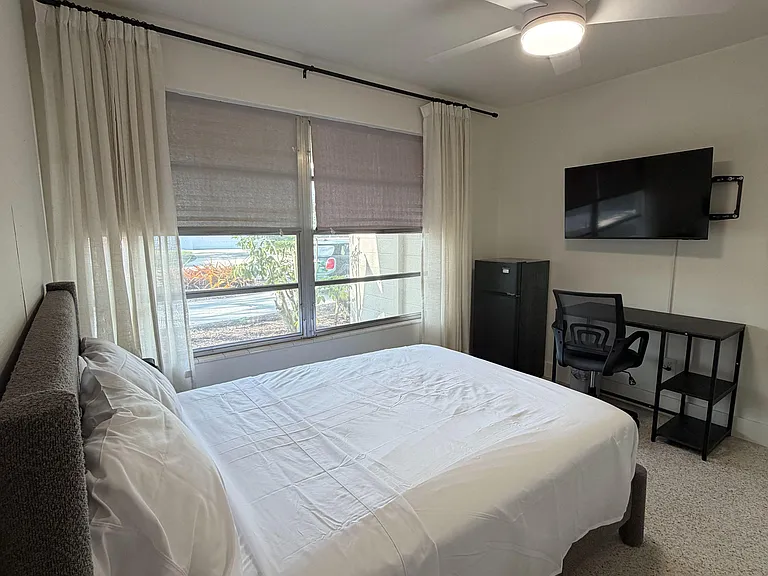 Furnished bedroom with a large window and layered curtains, plus a desk setup, mini fridge, wall-mounted TV, and ceiling fan.