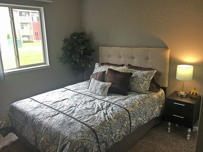 Bedroom with grey walls, tan carpet, queen bed, wooden nightstand with lamp to the right of the bed, and to the left of the bed is a large window.