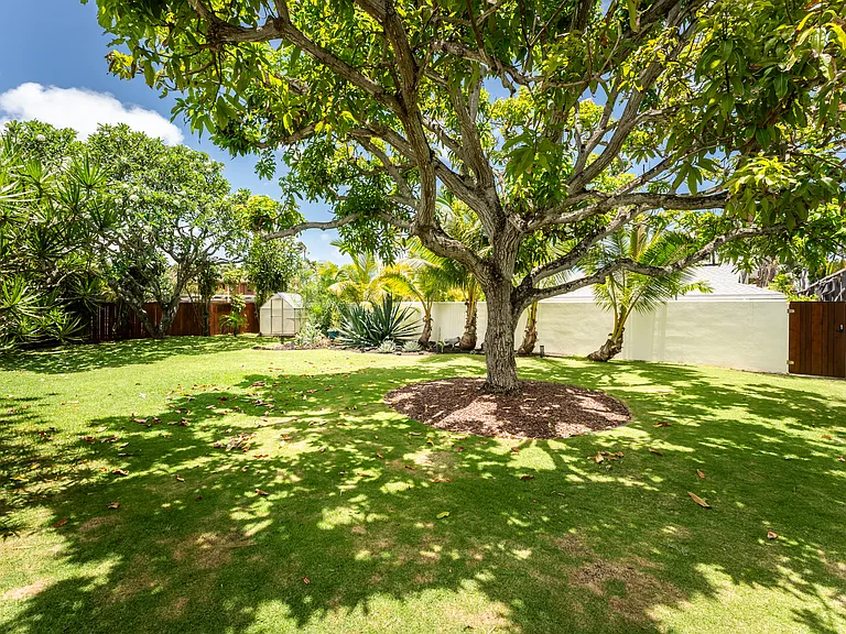 Landscaped backyard with mature shade trees, tropical greenery, and gated access to the private beach pathway.