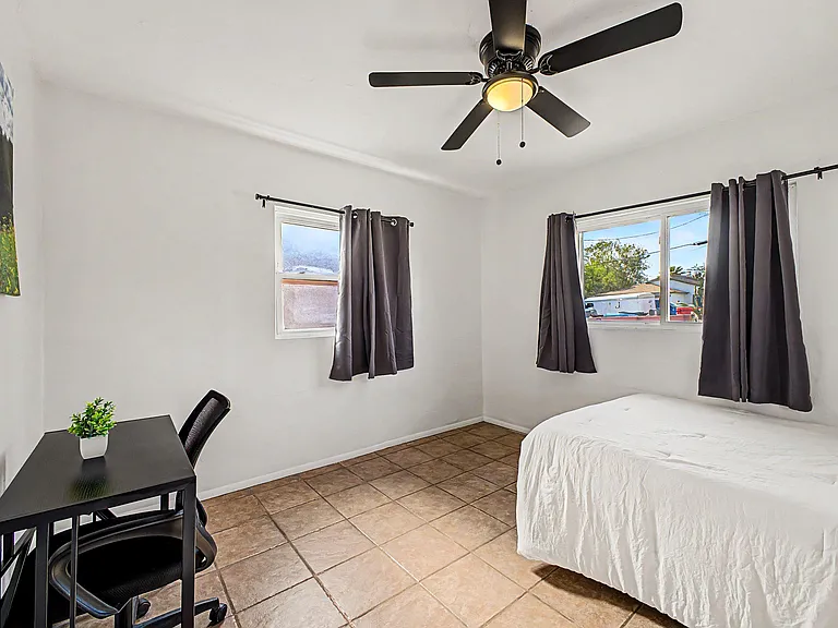 Bedroom with ceiling fan, tiled floors, two windows with blackout curtains, and space for a bed and compact desk.