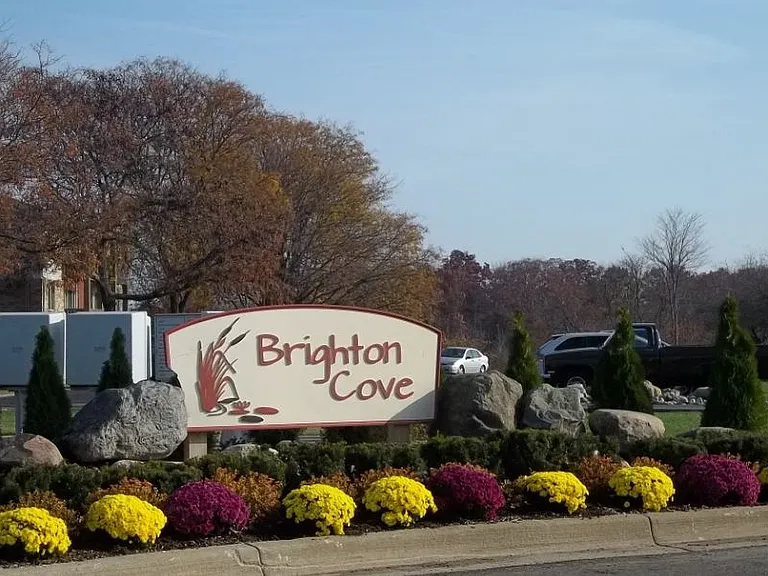 Beautifully landscaped monument sign at Brighton Cove. The sign is diffrent shades of brown. There are green hedgeds and red, and yelllow mums in front of the monument sign.