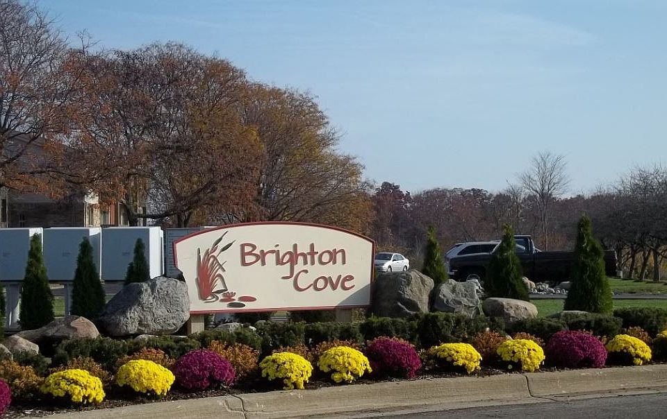 Beautifully landscaped monument sign at Brighton Cove. The sign is diffrent shades of brown. There are green hedgeds and red, and yelllow mums in front of the monument sign.