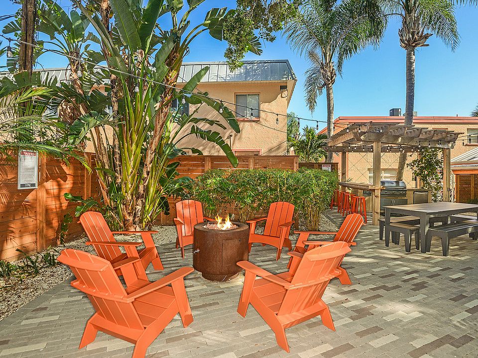 Inviting outdoor patio with vibrant orange chairs centered around a cozy fire pit, surrounded by lush greenery and a well-equipped barbecue area under a charming pergola.