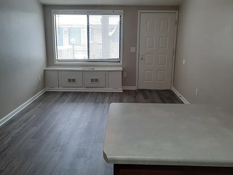 Living room in the Hampton floorplan. Picture is from the open kitchen showing the front window with light shining through. The floor is a gray VCT floor and there is a small ledge by the front window.