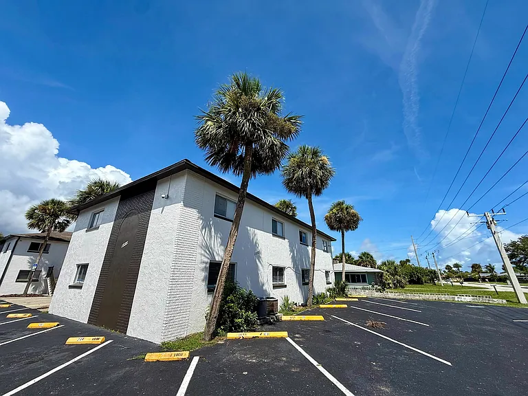 Charming apartment building with pristine white exterior and lush palm trees, offering ample parking and vibrant blue skies above.