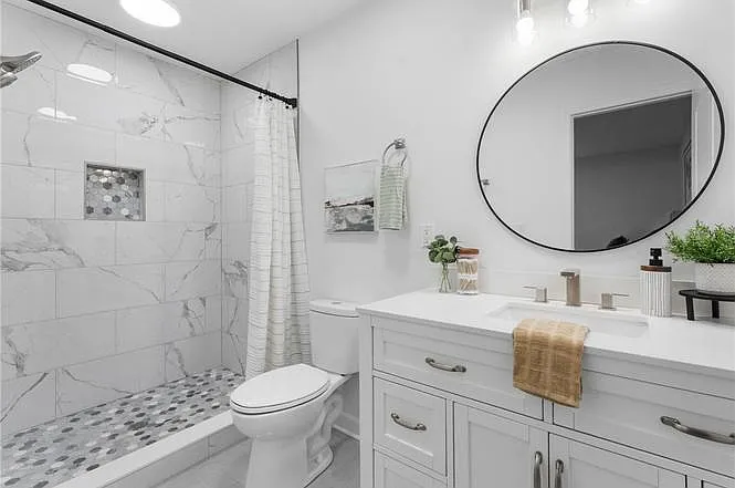 Updated full bath with large vanity, round mirror, and tiled tub/shower featuring a built-in niche and pebble-style floor.