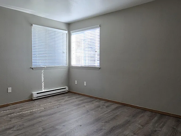 Cozy master bedroom with wood-style flooring, neutral walls, and two windows that fill the space with natural light. Features a clean, comfortable layout with baseboard heating and plenty of room to make it your own.