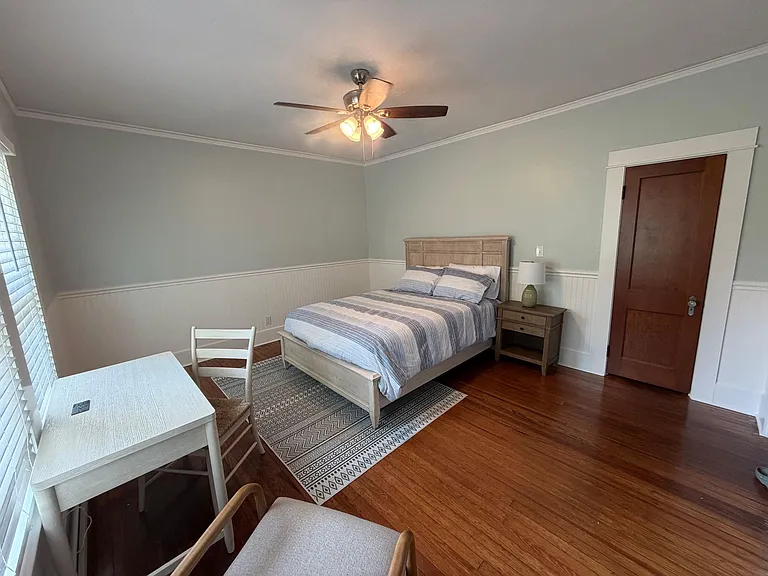 Furnished bedroom with hardwood floors, a ceiling fan, and a dedicated desk area, plus window blinds for light control.