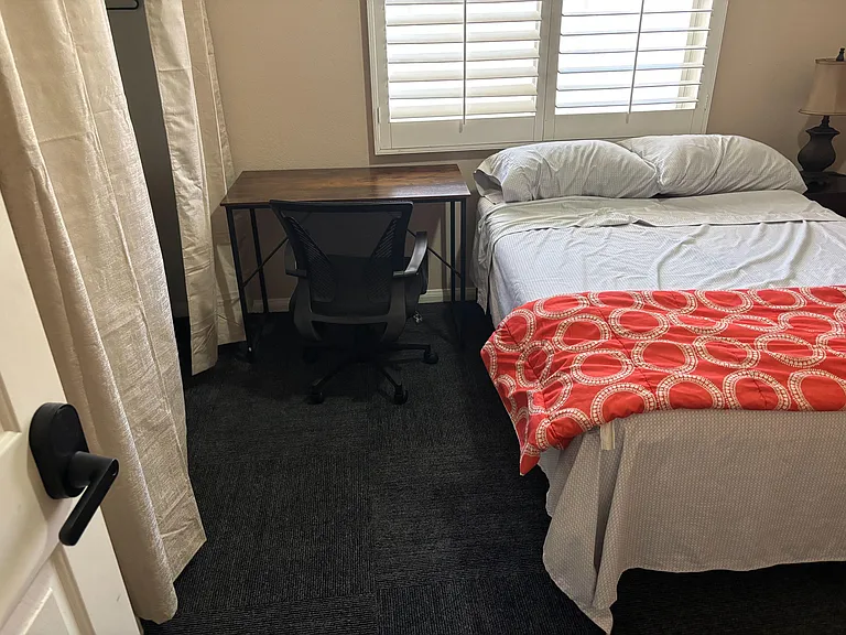 Furnished bedroom with a comfortable bed, dedicated desk workspace, and window shutters for light and privacy control.