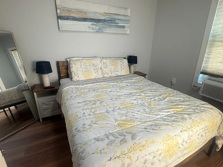 Furnished bedroom with a full bed, matching nightstands and lamps, and a window with blinds for light and privacy.