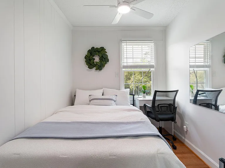 Furnished bedroom with a full bed, work desk, ceiling fan, and a large window with blinds for natural light.