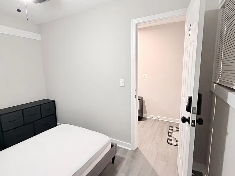 Furnished bedroom with a bed, dresser, ceiling fan, and easy access to the hallway; neutral finishes and window blinds.