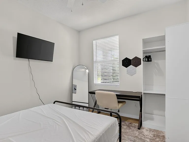 Furnished bedroom with a wall-mounted TV, work desk, and open closet storage, plus a window with blinds for privacy.