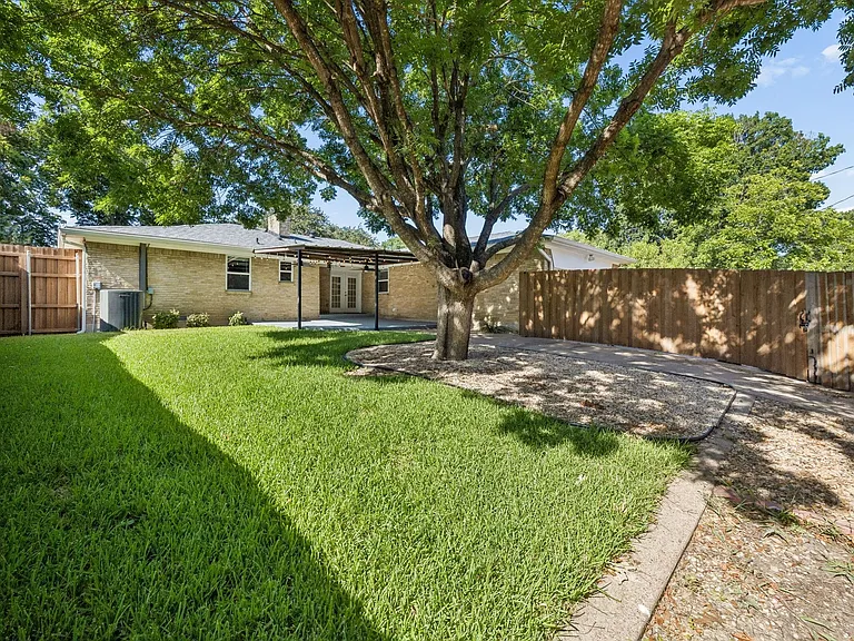 Spacious backyard featuring a large tree, manicured lawn, gravel area, privacy fencing, and a covered patio with a light-colored brick exterior