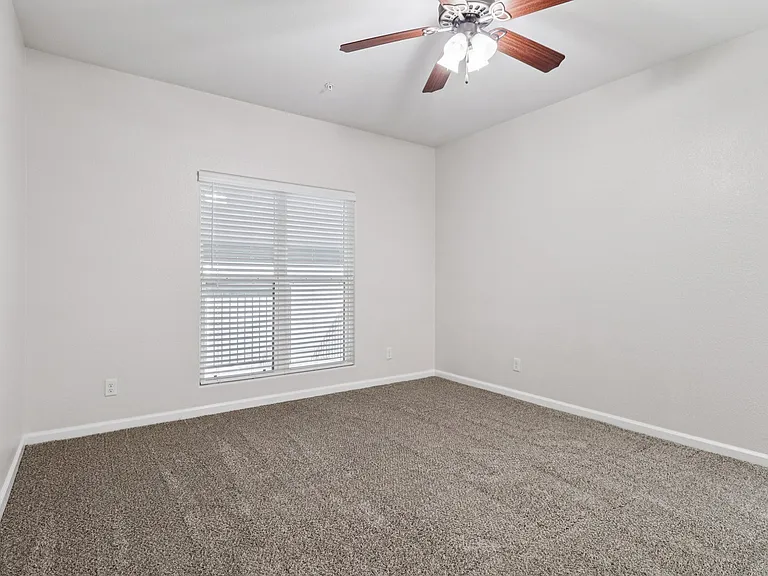 Bright, empty room with plush carpeting, a ceiling fan, and large window illuminating the space. Bright, empty room with plush carpeting, a ceiling fan, and large window illuminating the space.