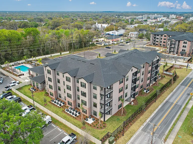 Bird's-eye view of a modern apartment complex featuring stylish architecture, lush greenery, ample parking, and a refreshing pool area, set in a vibrant neighborhood.