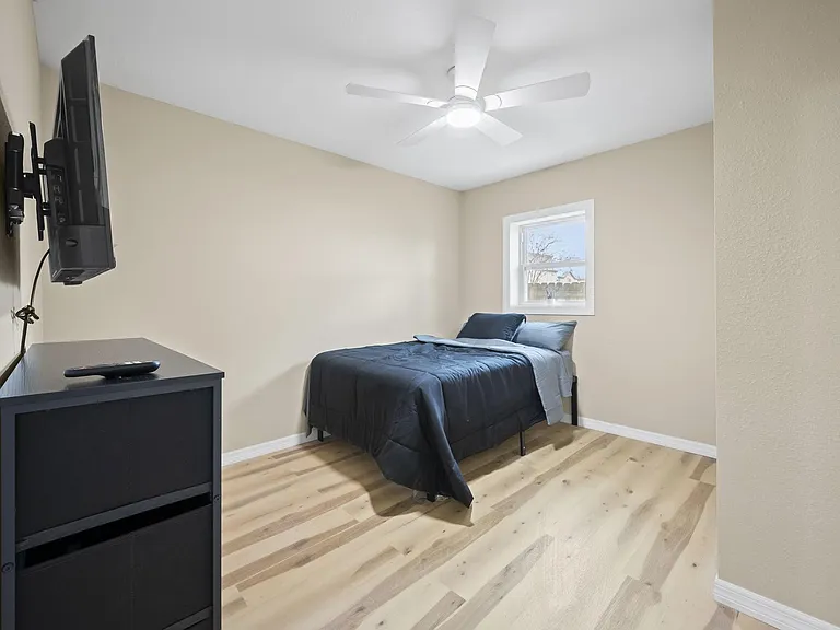 Compact bedroom with a ceiling fan, wood-look flooring, and a window for natural light; includes a bed and wall-mounted TV.