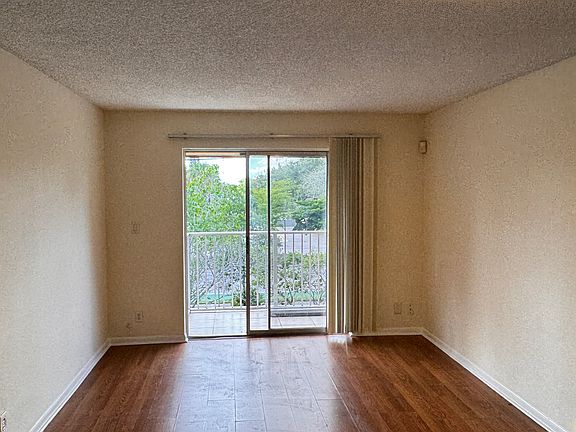 Bright and inviting living space featuring warm vinyl flooring and neutral walls, perfect for decorating. A large sliding glass door fills the room with natural light and opens to a private balcony with leafy views, creating an airy indoor-outdoor feel.