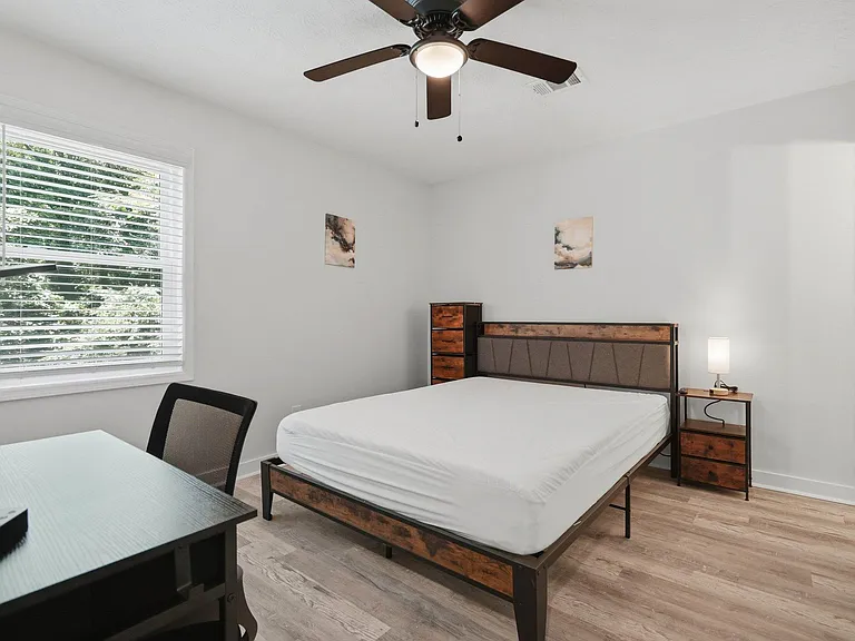 Private bedroom with a large window, ceiling fan, and space for a desk, furnished with a bed, nightstand, and dresser.