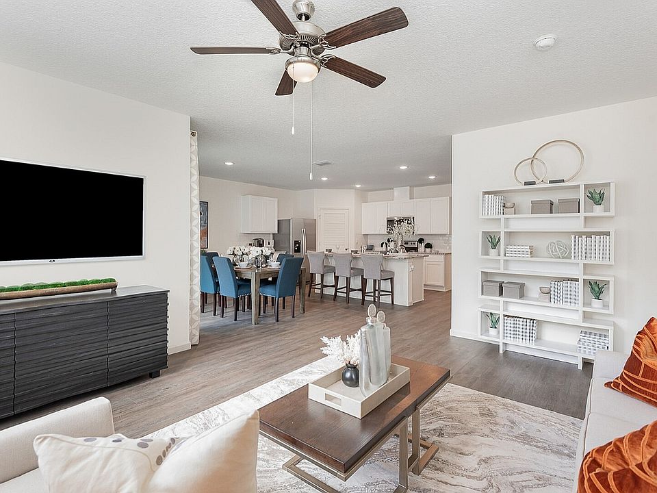Inviting open-concept living area featuring a modern kitchen, elegant dining space with blue chairs, and a cozy living room with stylish decor and a ceiling fan.