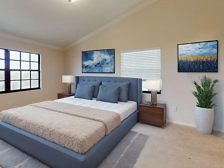Elegant bedroom with a plush blue bed, soft lighting, and large windows providing ample natural light. Stylish artwork and a potted plant add a fresh, contemporary touch.