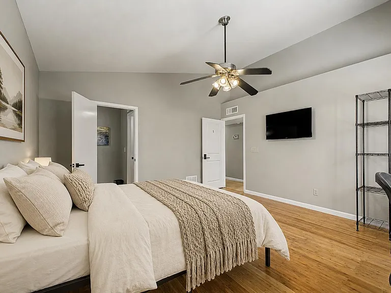Spacious bedroom with hardwood floors, ceiling fan, wall-mounted TV, and room for storage shelving and a work area.