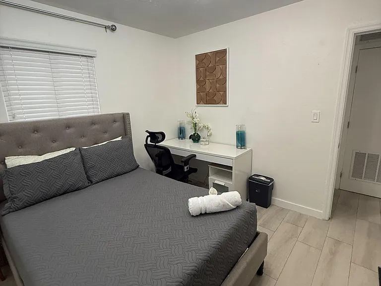 Furnished bedroom with a full bed, dedicated desk/workspace, and window with blinds for privacy and natural light.