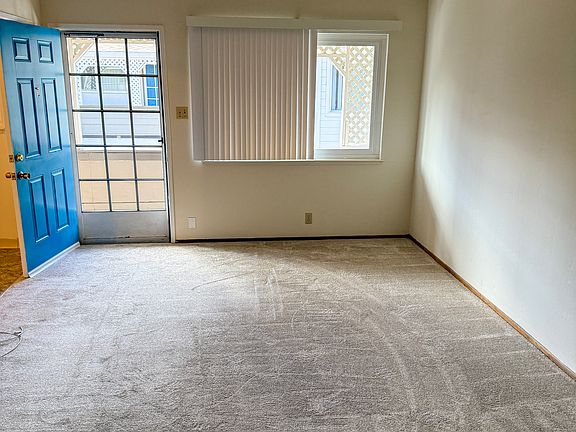 A bright, spacious living room featuring new carpeting and crisp white walls. The living space has a large window with vertical blinds provides plenty of natural light.