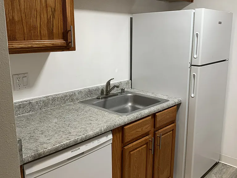 Compact kitchen featuring warm wood cabinetry, a neutral countertop, and easy-care wood-style flooring. Includes a stainless sink, dishwasher, and full-size refrigerator, with ample cabinet storage for everyday convenience.