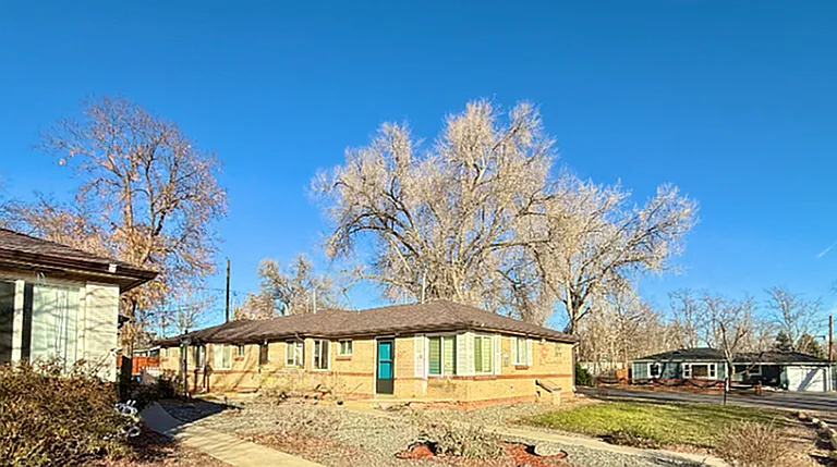 Charming single-story brick home with large windows, surrounded by mature trees and set against a bright blue sky.