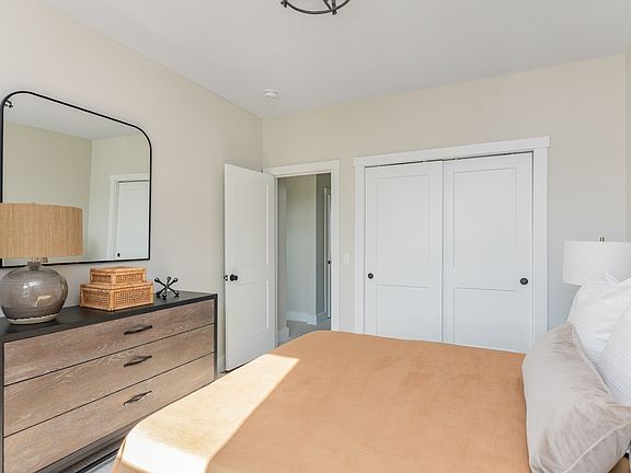 A well-lit, modern bedroom featuring a large bed, a dresser with a lamp and mirror, and white doors