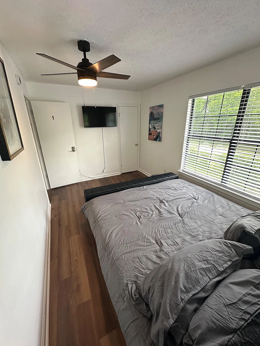 Bedroom features a ceiling fan with light, wall-mounted TV, and a large window with blinds plus wood-style flooring. - Avondale Cir