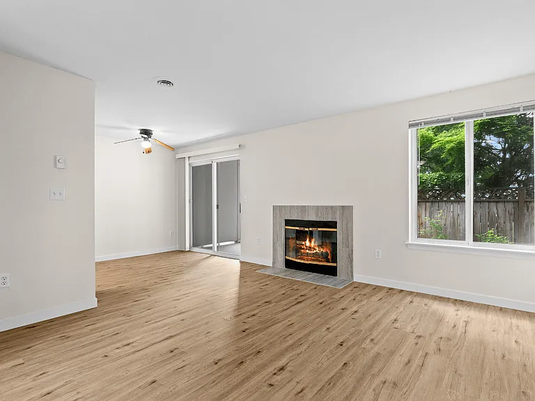 Spacious living room with wood-style plank flooring, cozy fireplace, ceiling fan, and sliding glass door patio access at Garden Square Apartments in Lakewood, WA. Spacious living room with wood-style plank flooring, cozy fireplace, ceiling fan, and slidin
