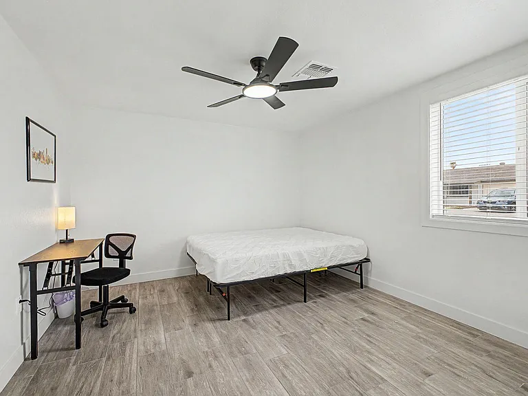 Private bedroom with easy-care flooring, ceiling fan with light, and a work desk beside a large window with blinds.