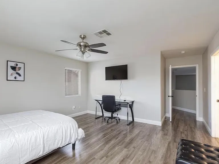 Spacious bedroom with wood-style flooring, ceiling fan, and a work desk setup, plus wall-mounted TV and window blinds.