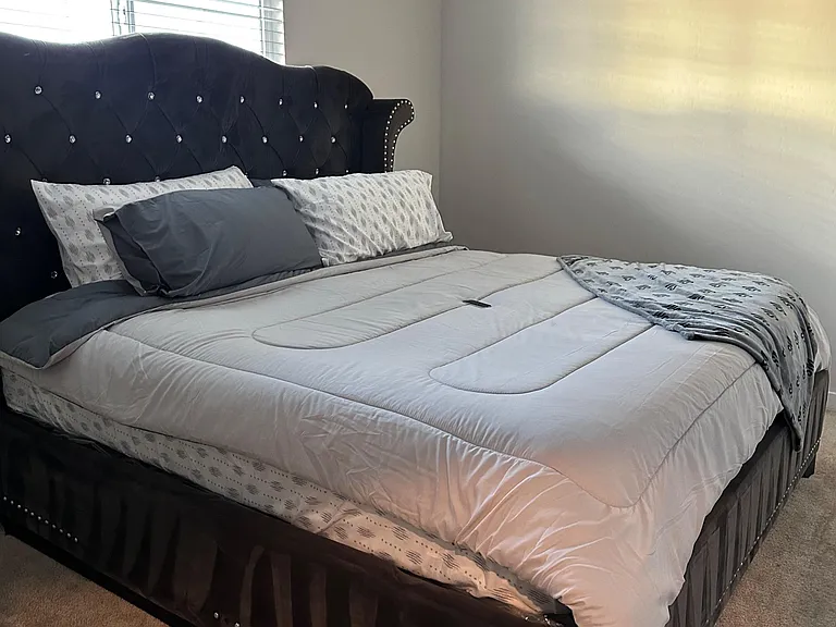 Furnished bedroom with a large bed, plush headboard, carpet flooring, and a window with blinds for light control.