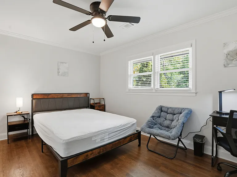 Spacious furnished bedroom with hardwood floors, ceiling fan, large window, and a dedicated desk area for working.