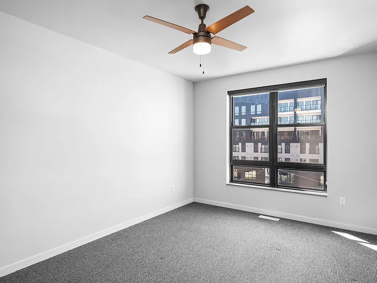 Bright empty room featuring modern ceiling fan, large window with city view, and cozy carpet flooring. Bright empty room featuring modern ceiling fan, large window with city view, and cozy carpet flooring.