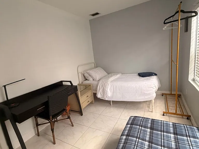 Private bedroom with tiled floors, a work desk, and a sunny window with blinds plus an open garment rack for storage.