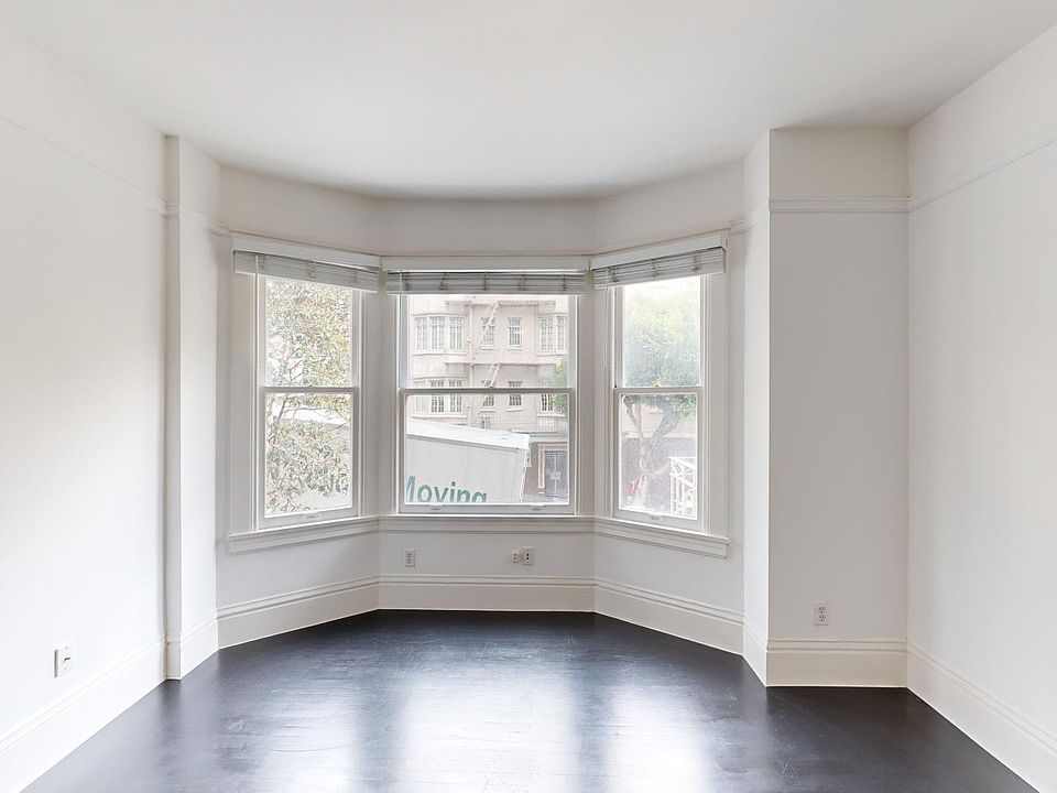 Empty room with hardwood floors, walls, and a large bay window with blinds. Outside, a moving truck is partially visible. Bright and open atmosphere.