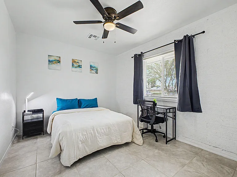 Furnished bedroom with a ceiling fan, large window with curtains, and a dedicated desk workspace beside the bed.
